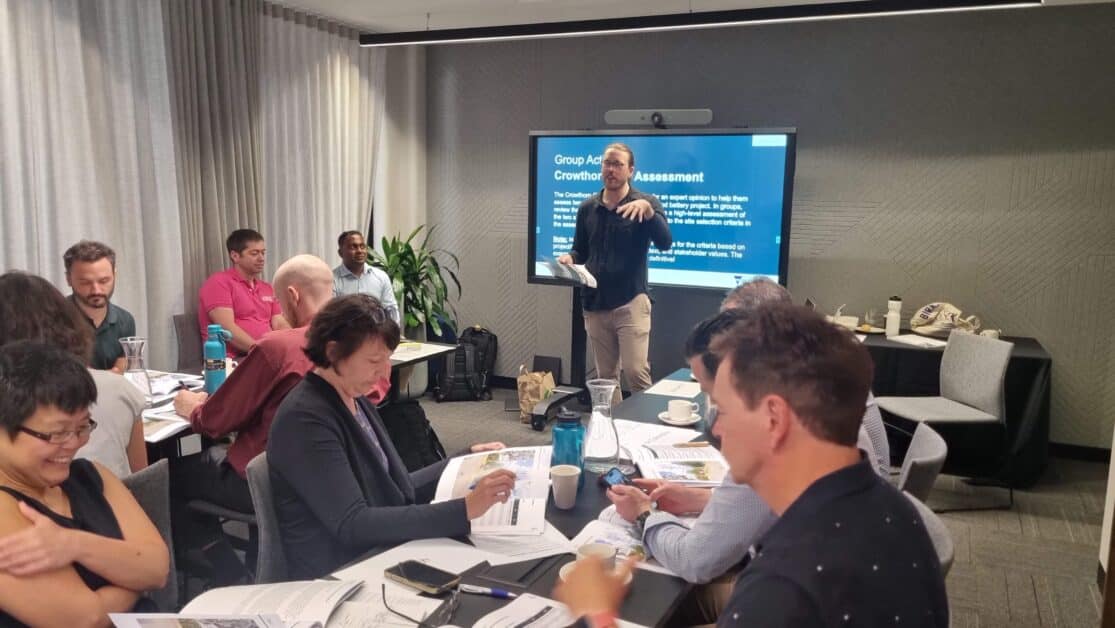 A group of participants are engaged in a discussion during a group activity in a conference room. A presenter stands at the front, gesturing while speaking, with a large screen displaying a presentation behind him. Attendees are seated at tables, reviewing documents and taking notes. The room is well-lit with large windows and curtains.