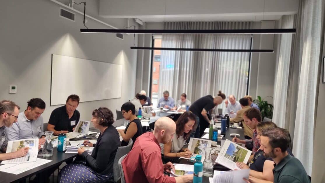 A group of people are seated around tables in a well-lit conference room, engaged in discussions and reviewing documents. The room has large windows with curtains, and there are multiple small groups working together, each appearing focused on their tasks. Various materials and water bottles are visible on the tables.