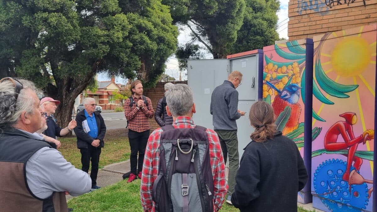 A group of people are gathered outdoors around a neighbourhood battery with mural art, which includes images of a bird, flowers, and a person riding a bicycle. One person is speaking to the group while others listen attentively. Trees and residential buildings are visible in the background.