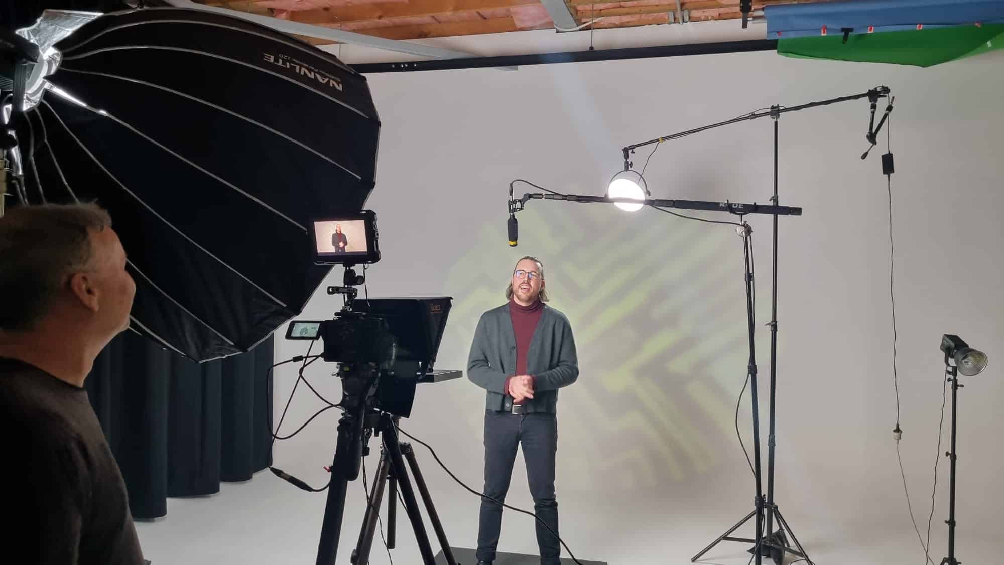 A presenter stands in front of a camera and professional lighting setup during a video shoot. The presenter, wearing glasses, a maroon turtleneck, and a grey cardigan, speaks in front of a white backdrop with a projected geometric pattern. Microphones are suspended above the presenter, and a person monitors the scene behind a camera rig equipped with a monitor displaying the presenter.