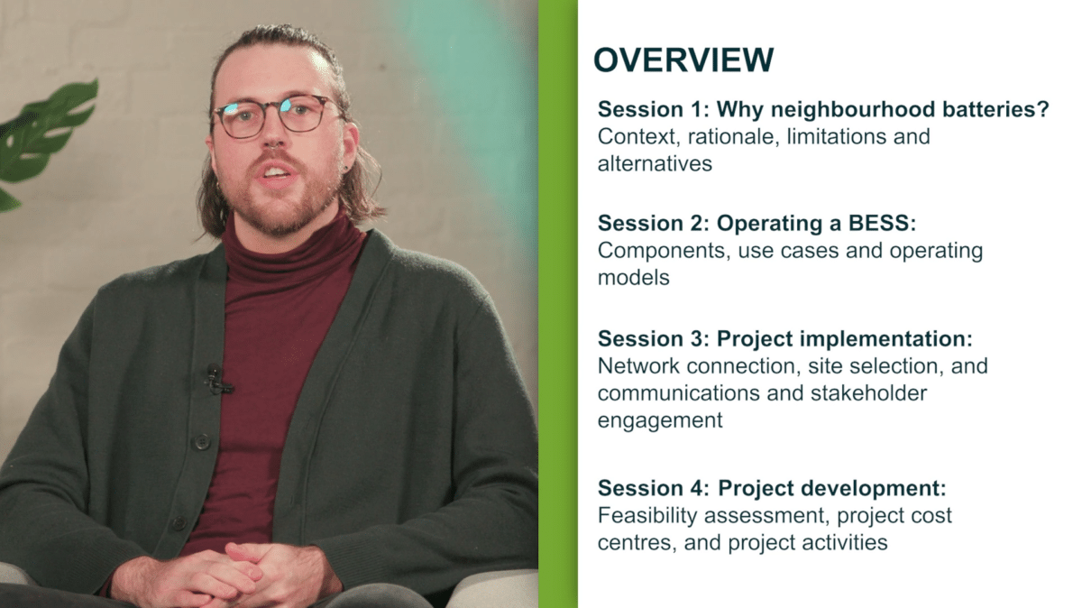 A man wearing glasses, a maroon turtleneck, and a dark green cardigan is speaking on camera. Next to him, a slide titled "Overview" is displayed, outlining four sessions: Why neighbourhood batteries? - Context, rationale, limitations, and alternatives. Operating a BESS: - Components, use cases, and operating models. Project implementation: - Network connection, site selection, communications, and stakeholder engagement. Project development: - Feasibility assessment, project cost centres, and project activities. A potted plant is visible in the background, and the overall setting is professional and informative.